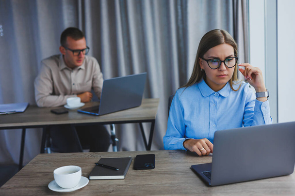 İş arkadaşları modern geniş bir ofiste arka planda çalışırken, günlük kıyafetleriyle bir masada oturup dijital tablet kullanarak kahkaha atan genç bir işadamı.. - Fotoğraf, Görsel