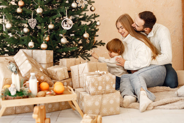 Daddy and mom with their son near christmas tree with lights and gifts hugging and having fun. Cheerful family celebrating Christmas. Mother, father and little boy wearing white sweaters. - Fotoğraf, Görsel