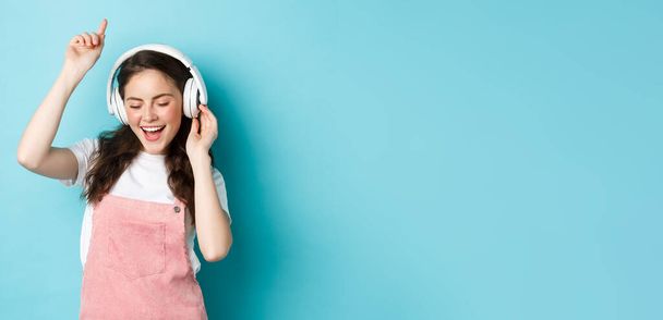 Stílusos Y-generációs lány táncol tavaszi ruhában, zenét hallgat fejhallgatóban és élvezi a hangot, áll a kék háttér felett. - Fotó, kép