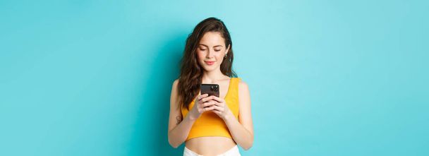 Stilvolle junge Frau checkt Nachrichten am Telefon, liest Smartphone-Bildschirm und lächelt, shoppt online, steht vor blauem Hintergrund. - Foto, Bild