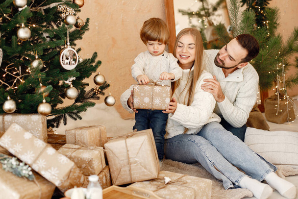 Daddy and mom with their son near christmas tree holding a gift. Cheerful family celebrating Christmas. Mother, father and little boy wearing white sweaters. - Фото, изображение