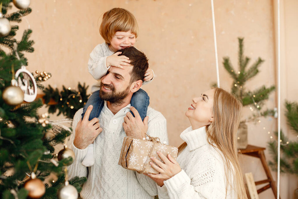 A daddy and mom with their son decorating christmas tree with toys. Son sitting on fathers shoulders. Mother, father and little boy wearing white sweaters. - 写真・画像