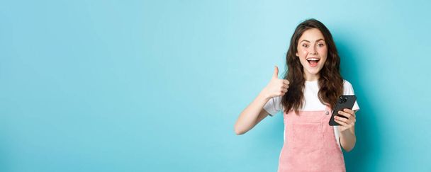 Portrait of excited cute girl say yes, smiling and nod in approval, holding smartphone while showing thumb up, like and praise good online offer, standing against blue background. - Fotografie, Obrázek