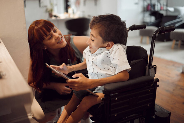 Mujer, discapacidad infantil y tableta, silla de ruedas amigable e inclusiva, necesidades especiales y apoyo. Parálisis cerebral y discapacidad infantil, tiempo de pantalla y juegos, discapacitados en el salón de peluquería - Foto, imagen
