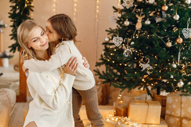 Portrait of a little girl celebrating Christmas and hugging. Girl and her mother sitting near Christmas tree and posing for a photo. Family wearing beige knitted clothes. - Fotó, kép