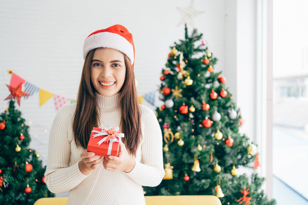 Young Asian woman wearing a sweater and Santa Claus hat smiles and holds a Christmas gift in a red box with ribbon in her hands a living room with a decorated Christmas tree in the background. - Foto, imagen