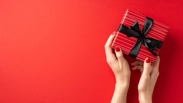 Black friday concept. First person top view photo of girl's hands holding red giftbox with black ribbon bow on isolated red background with copyspace - Photo, image
