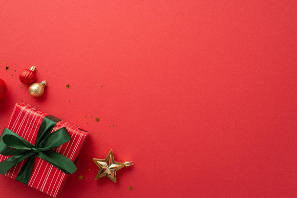 Concept de Noël. Photo de vue du dessus de boules or étoile ornement boîte-cadeau rouge avec ruban vert arc et confettis sur fond rouge isolé avec espace vide - Photo, image