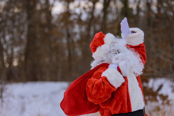 Белый снежный вид окружает Санта-Клауса ходить рядом с лесом, держа красные мешки заполнены подарками для детей на Рождество - Фото, изображение