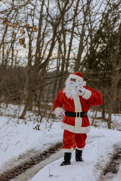 Santa Claus is walking near forest carrying red bag full of presents for children around white background covered in snow - Φωτογραφία, εικόνα