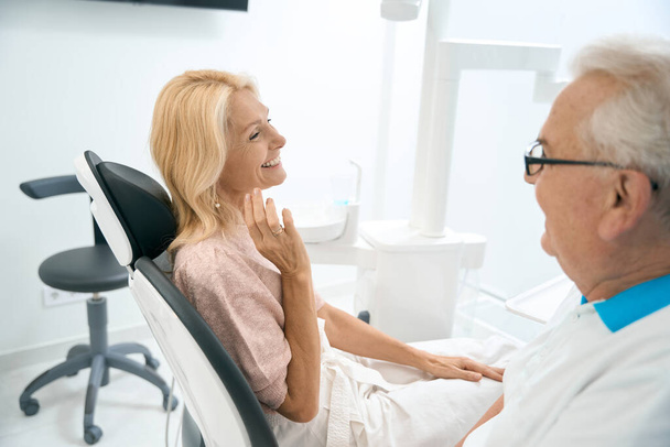 Pas w górę widok z boku portret uśmiechniętej pięknej pani siedzi w fotelu dentysty podczas dyskusji z dentystą w centrum medycyny - Zdjęcie, obraz