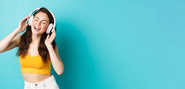 Jeune femme en vêtements d'été écoutant de la musique, portant des écouteurs et chantant le long de la chanson préférée, dansant dans des écouteurs, debout sur fond bleu. - Photo, image