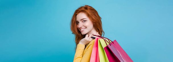 ショッピングのコンセプト - ショッピング バッグとカメラを見て笑っている肖像若い美しい魅力的な redhair 女の子をクローズ アップ。青いパステル背景。コピー スペース. - 写真・画像