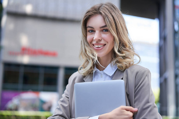 Retrato de jovem mulher corporativa, segurando seu laptop e sorrindo, indo para o trabalho, vestindo roupa profissional, suíte bege. - Foto, Imagem