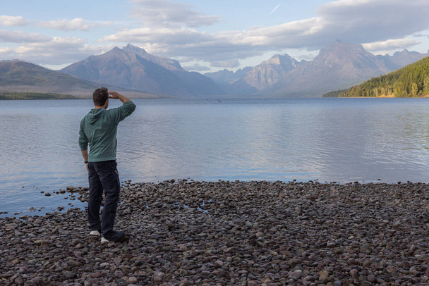 Man standing on the shoreline looking at the amazing mountains across lake McDonald in Glacier National Park - Photo, Image