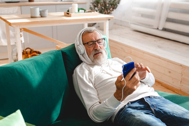 Hombre hipster senior que usa el teléfono mientras escucha música de lista de reproducción en la aplicación móvil: hombre mayor que usa máscara protectora para la cara mientras ve el teléfono inteligente de vídeo - Foto, imagen