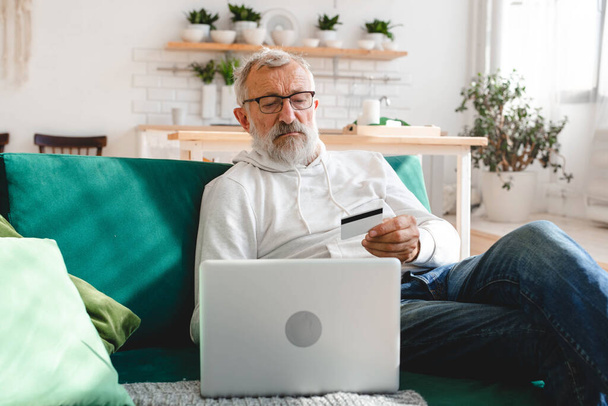Idősebb férfi, aki online fizet laptoppal. Mosolyogni az öreg nagyapának, miközben e-kereskedelemben vásárol hitelkártyával. Az internetes banki betéti kártyával rendelkező nyugdíjas - Fotó, kép