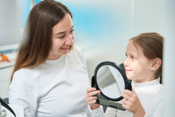 Pleased pre-teen child with mirror in hands looking at cheerful pediatric dentist - Foto, Imagen