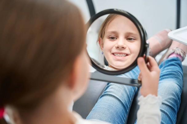 Concentrated little patient sitting in dental chair and looking at her open mouth in hand mirror - Fotoğraf, Görsel