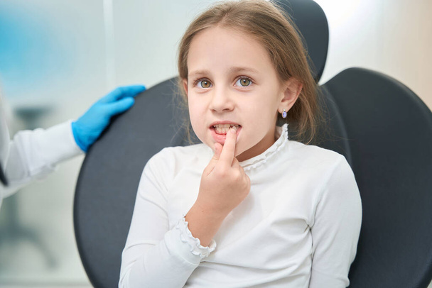 Pre-teen girl seated in dentist office pointing at problem tooth in her mouth - Photo, Image