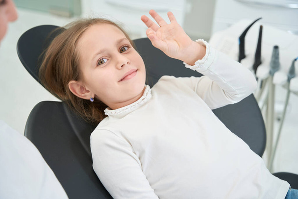 Pleased preteen girl lying in dental chair and making OK sign before camera - 写真・画像