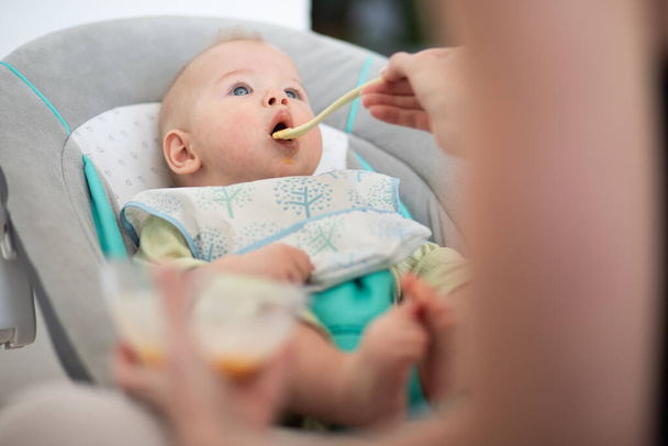 Cucchiaio madre nutrire il suo bambino bambino sulla sedia del bambino con purea di frutta. Concetto di introduzione di alimenti solidi per bambini - Foto, immagini