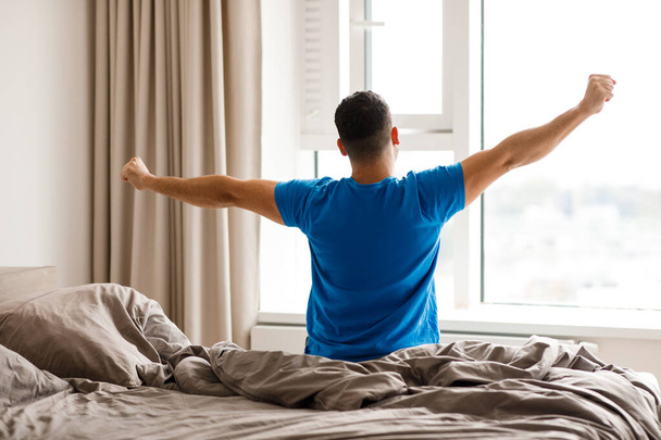 Een man wordt wakker in een slaapkamer tegenover het raam en steekt zijn handen uit. Hoge kwaliteit foto - Foto, afbeelding