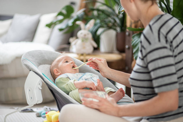 Mère cuillère nourrir son bébé garçon enfant dans une chaise bébé avec de la purée de fruits. Concept d'introduction aux aliments solides pour bébés - Photo, image