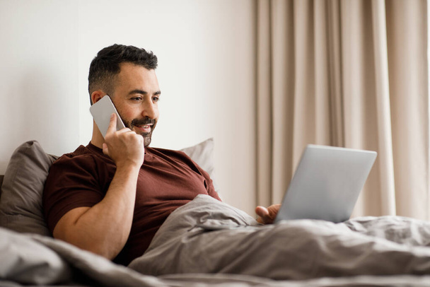 Emocionado hombre europeo acostado en la cama, hablando por teléfono, trabajando en un ordenador portátil, espacio libre para copiar. Foto de alta calidad - Foto, imagen