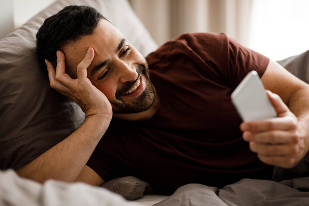 Счастливый улыбчивый молодой парень постукивает по экрану телефона в постели. Высокое качество фото - Фото, изображение