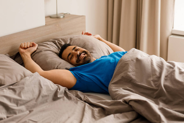 Радостный европейский мужчина тысячелетия улыбается с закрытыми глазами, лежит в постели в пижаме, расслабляется в современной спальне дома. Высокое качество фото - Фото, изображение