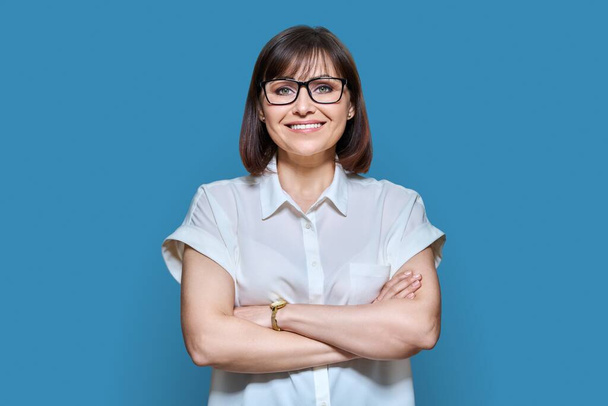 Attractive confident middle aged business woman looking at camera on blue color background. Smiling 40s female with glasses, crossed arms. Lifestyle, confidence, work job business, mature people - Fotografie, Obrázek