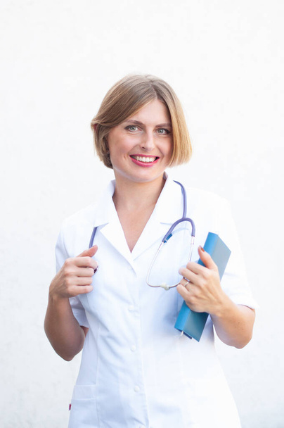 Young beautiful caucasian woman doctor with a phonendoscope on her neck and with a notepad smiling on a light background. High quality photo - Foto, Bild