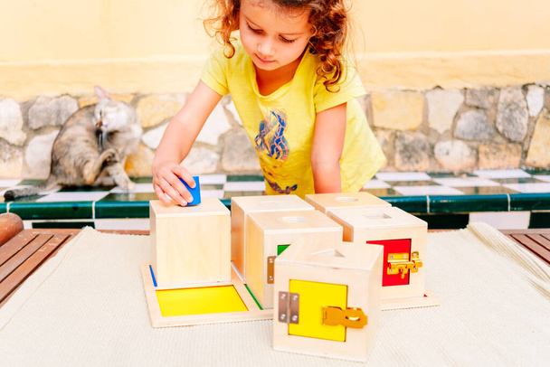 Ένα κορίτσι χειρίζεται αισθητηριακό υλικό montessori, ξύλινα κουτιά για να χωρέσει κύβους. - Φωτογραφία, εικόνα
