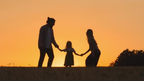 Three silhouettes happy carefree cheerful family parents mother father daughter woman and man with child girl kid in sunset sun jumping in wheat yellow golden field outdoors playful game jump in air - Footage, Video