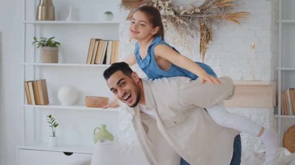 Funny Hispanic Indian man loving father dad holds on back little daughter kid plays with girl child piggyback airplane fly game baby holding arms to sides having fun at home pretend flying playing - Footage, Video
