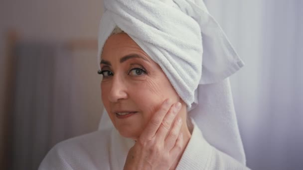 Close up female portrait happy 50s middle-aged lady 60s mature woman touching facial skin looking at camera with smile touch face pampering wears towel on head enjoy cosmetics and plastic surgery - Footage, Video