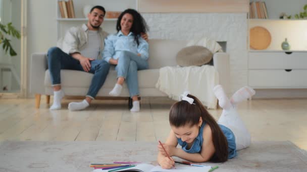 Küçük beyaz kız öğrenci kız öğrenci yere uzanmış renkli kalemlerle resim çiziyor hobi olarak çok ırklı ebeveynler anne ve baba kanepeye oturuyor. - Video, Çekim