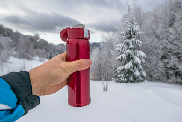 Red thermo bottle for winter leisure. Flask thermos of drinks in hand on background of winter mountains landscape - Photo, Image