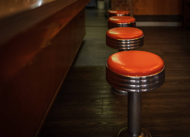 Stare stołki w restauracji. Wiersz vintage stołki przed drewnianą ladą wewnątrz vintage bar stylu. Rząd czterech błyszczących czerwonych stołków winylowych. Nikt, selektywne skupienie - Zdjęcie, obraz
