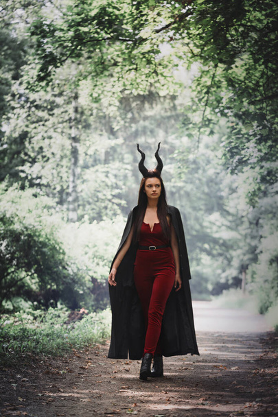 An incredibly beautiful witch in a black cloak with horns Maleficent walks through the dense forest. Halloween concept - Photo, Image