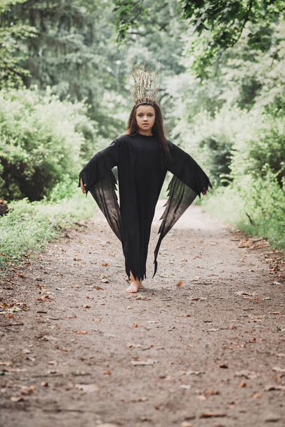 Młoda kobieta w czarnej sukience i koronie gałęzi spaceruje w ciemnościach lasu. Koncepcja Halloween - Zdjęcie, obraz