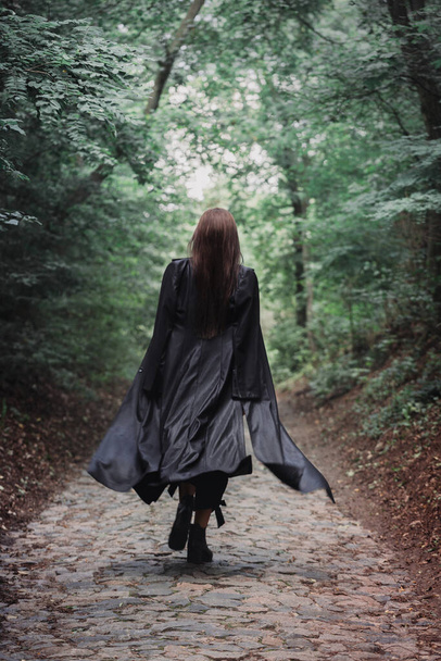 A tall beautiful woman in a black raincoat walks through a dense forest, back view. Halloween concept - Φωτογραφία, εικόνα