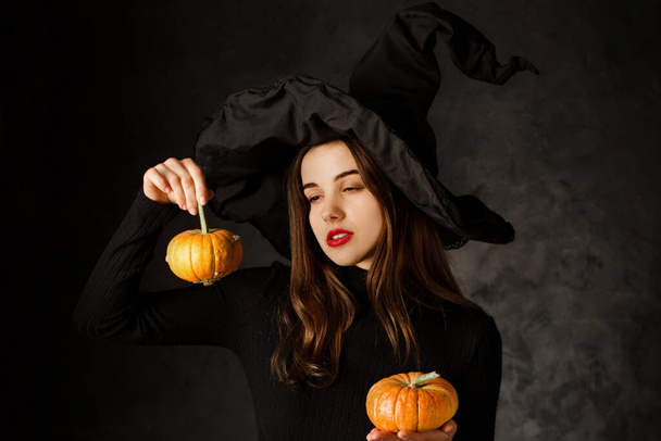jonge mooie vrouw in een zwarte heks hoed met kleine pompoenen op een zwarte achtergrond - Foto, afbeelding
