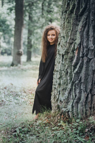 Cadılar Bayramı konsepti olan, ağacın yanındaki ormanda cadı kostümü giymiş küçük bir kız. - Fotoğraf, Görsel