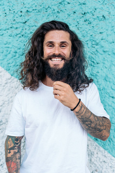 Cool bearded man with long hair smiling and looking at camera against blue wall. - Photo, Image