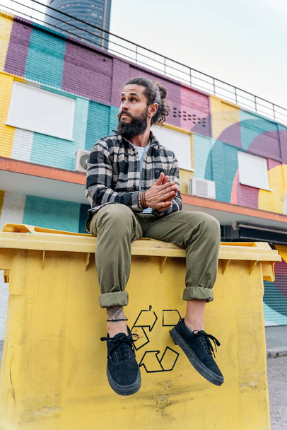 Cool hipster man with long beard sitting in a recycling container and looking to the side. - 写真・画像