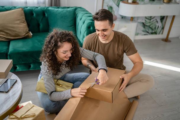 Ζευγάρι άντρας και γυναίκα ετοιμάζουν δώρα για διακοπές καυκάσιος άνδρας και γυναίκα συσκευασίας δώρα για τους φίλους χρησιμοποιώντας χαρτί περιτύλιξης χαρούμενο χαμόγελο κρατήστε το κουτί δώρου στο σπίτι πραγματικοί άνθρωποι αντιγράφουν χώρο - Φωτογραφία, εικόνα