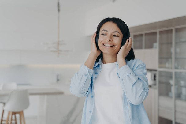 Lächelnde junge Frau, die mit drahtlosen Kopfhörern der Musik-Playlist lauscht und den Klang der Musik genießt. Glückliche Frauen hören Podcast, Hörbuch oder positive Audio-Affirmationen und ruhen sich zu Hause aus.  - Foto, Bild