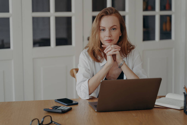 Porträt einer seriösen Managerin, die am Laptop am Schreibtisch sitzt und in die Kamera blickt. Selbstbewusste nachdenkliche junge Geschäftsfrau nutzt Computer für Remote-Job posiert am Arbeitsplatz.  - Foto, Bild